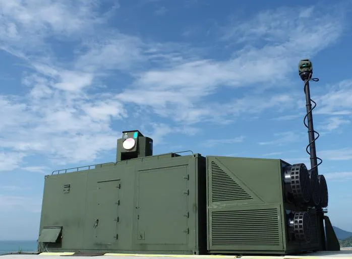A green containerised laser weapon system with a turret-mounted beam director and a mast-mounted sensor under a blue sky
