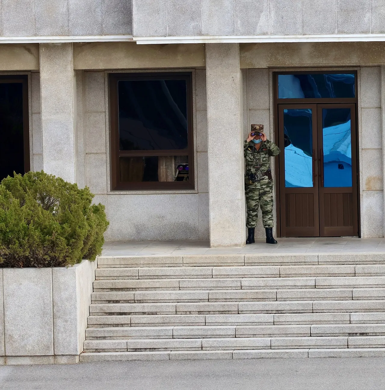 A North Korean soldier in camouflage uniform and cap stands by a glass door at the top of stone steps, looking through binoculars