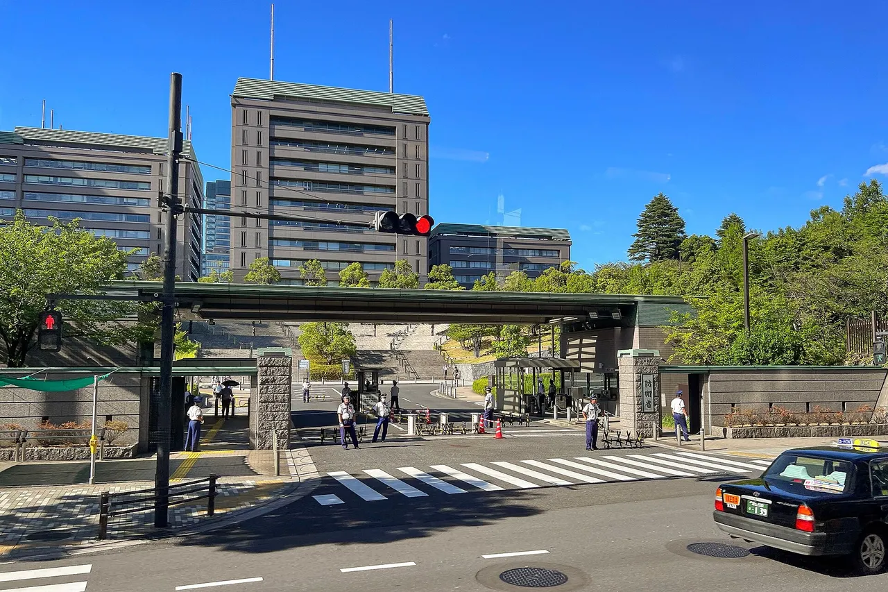 Gate and office towers of Japan's Ministry of Defense headquarters in Tokyo under a clear blue sky, with guards at the entrance