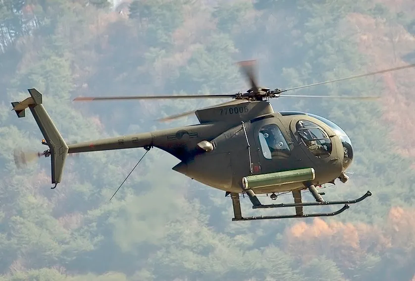 An olive-green MD 500 light military helicopter with a rounded glass cockpit flies low in front of a wooded hillside