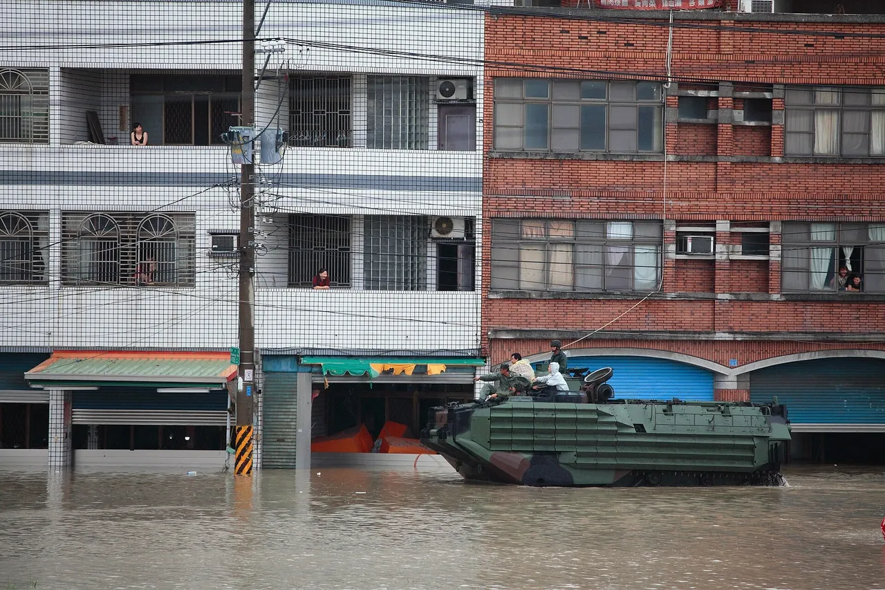 A green tracked amphibious assault vehicle with crew on top driving through a flooded street lined with shops and apartment blocks