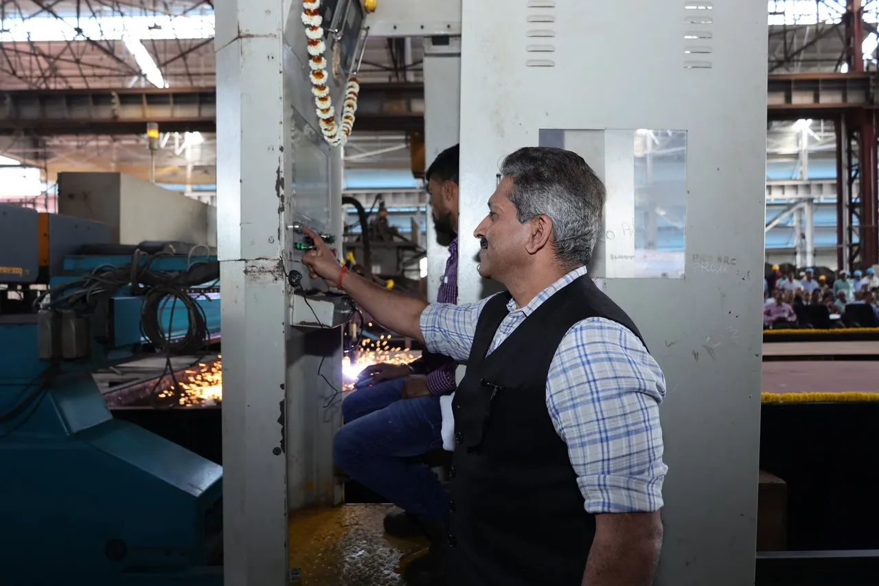 A man in a checked shirt and dark vest presses a button on a steel plate-cutting machine as sparks fly inside a large shipyard workshop
