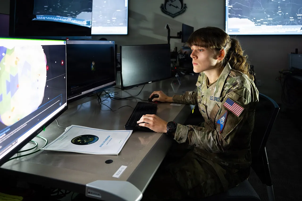 A uniformed US Space Force member sits at a desk with several monitors showing orbital and map displays in a darkened operations room