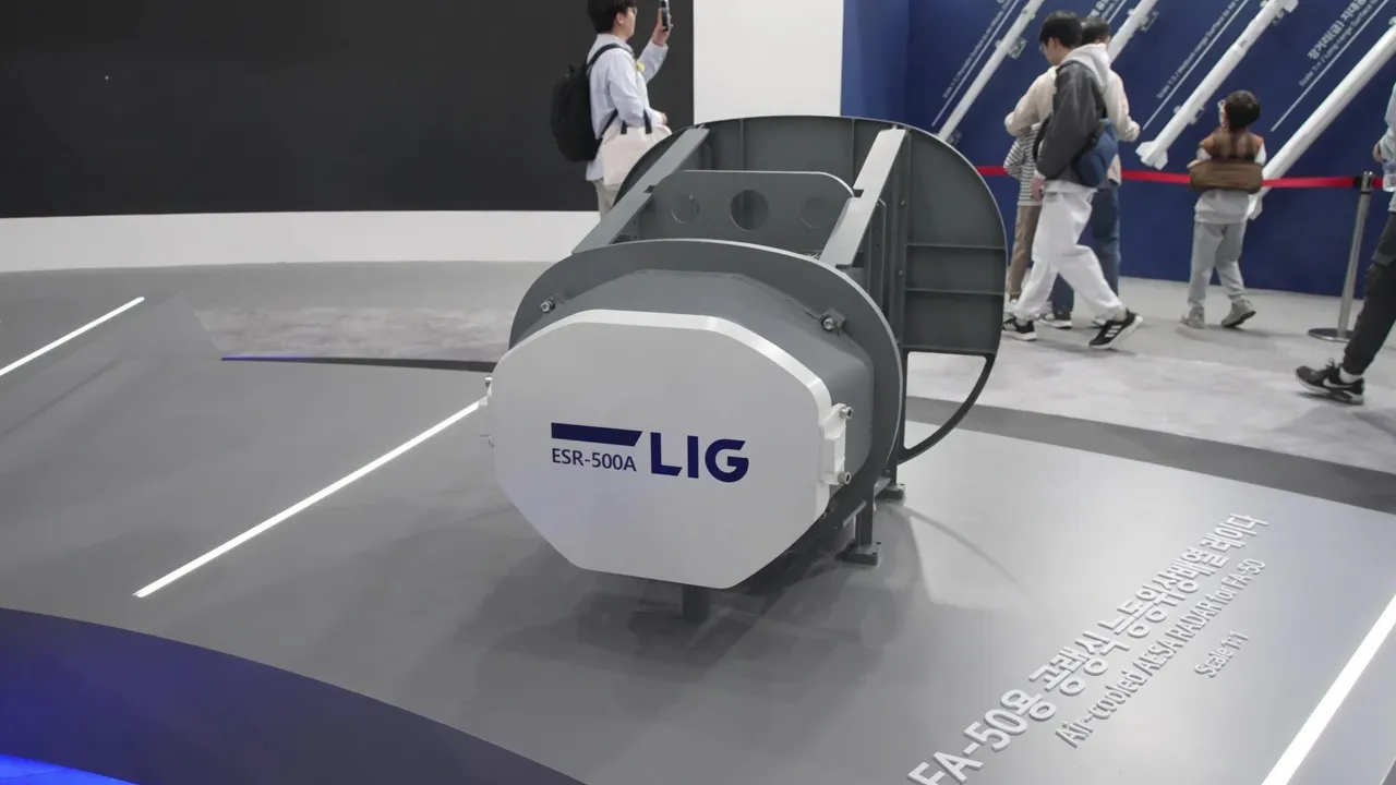 A grey and white boxy radar unit marked ESR-500A and LIG sits on a display stand at an exhibition while visitors walk past in the background