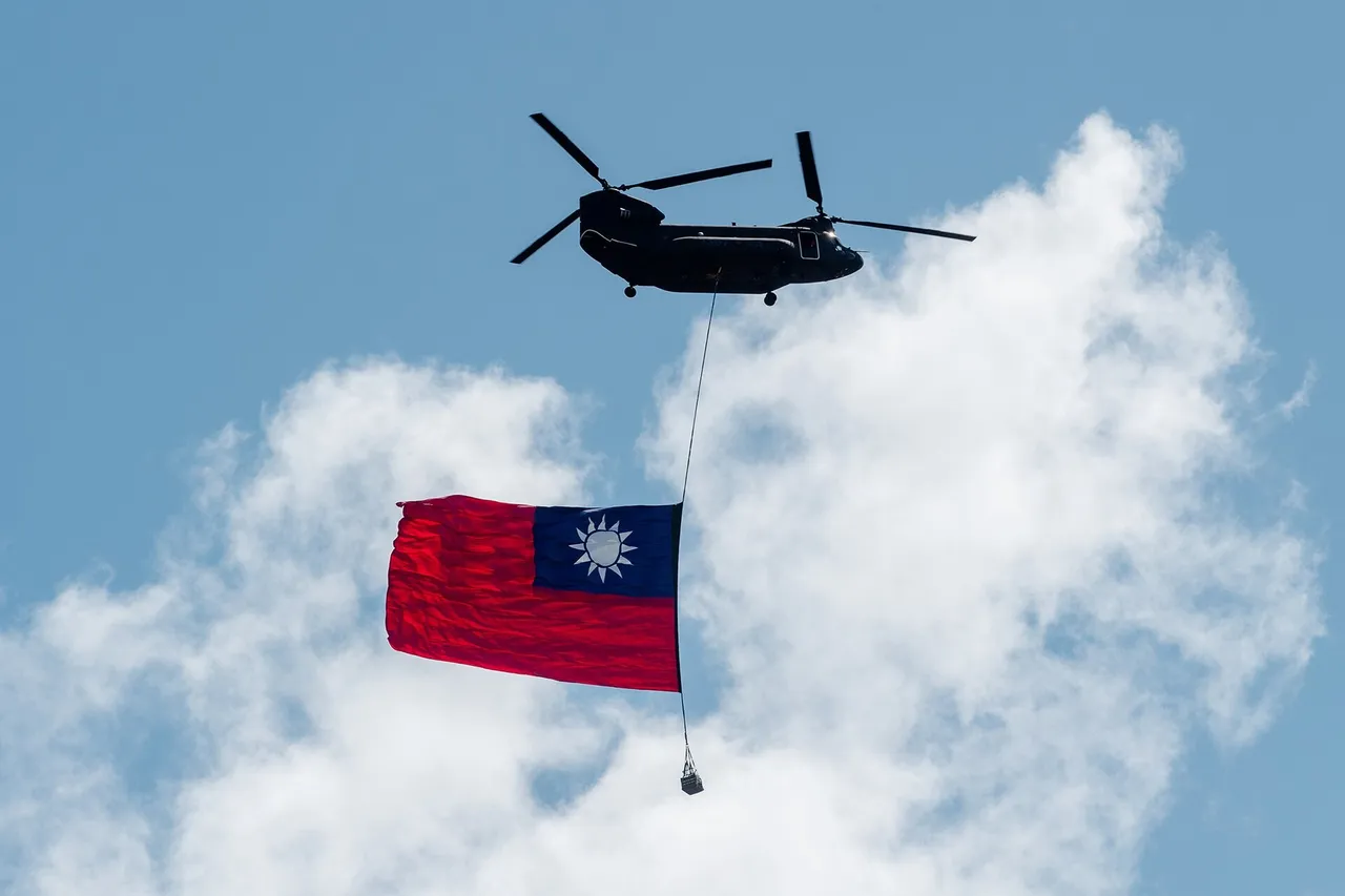 A dark twin-rotor Chinook helicopter flies against a blue sky with white clouds, a large red, blue and white Republic of China flag hanging beneath it on a long cable