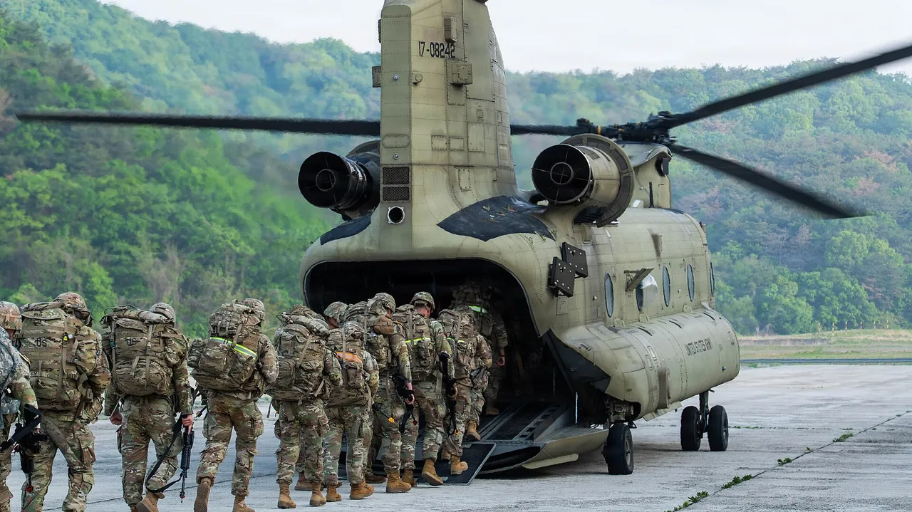 Soldiers in camouflage with backpacks and rifles file up the rear ramp of a tan CH-47 Chinook helicopter on a tarmac surrounded by green hills