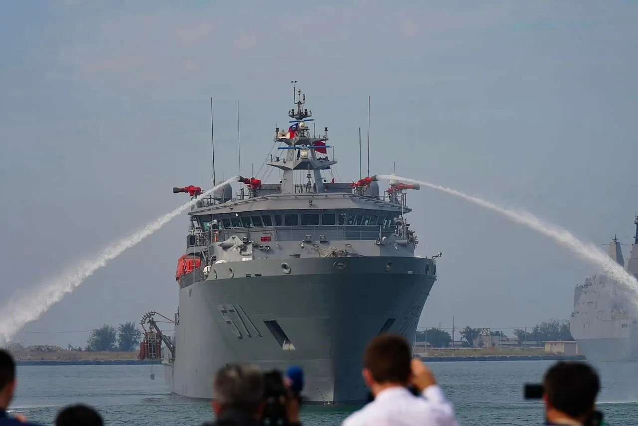 A grey navy rescue and salvage ship with a tall white superstructure and a large crane at the stern sailing on calm water.