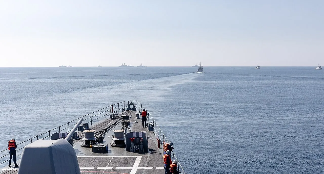 View from the bow of a grey warship, with crew in orange life vests on deck and several naval vessels spread across a calm sea ahead