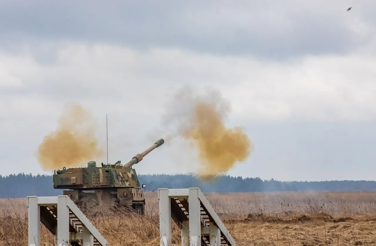 A camouflaged tracked howitzer fires its long barrel across a brown field, sending clouds of orange smoke into a grey sky