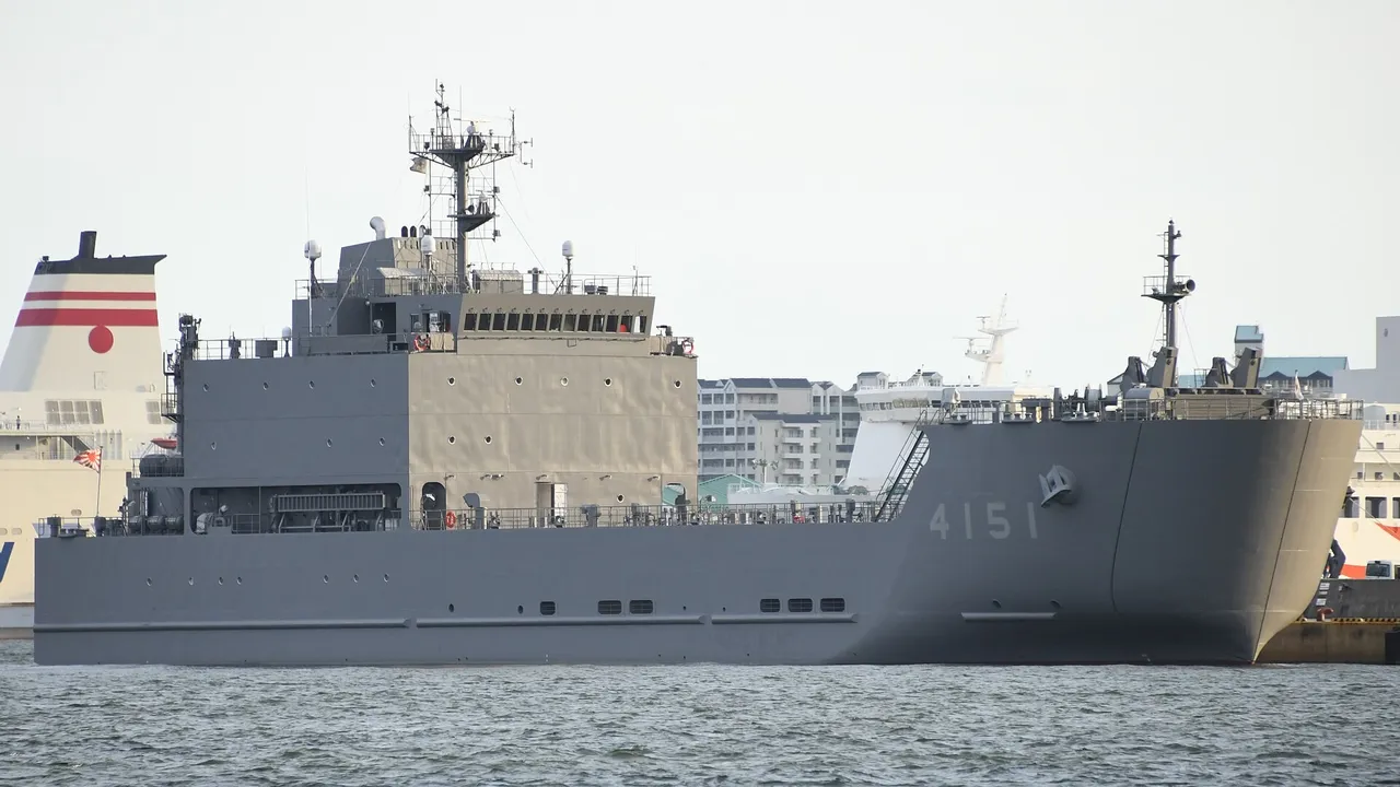 A grey flat-bowed military transport ship with hull number 4151 moored in a harbour, with buildings and a ferry funnel behind it.