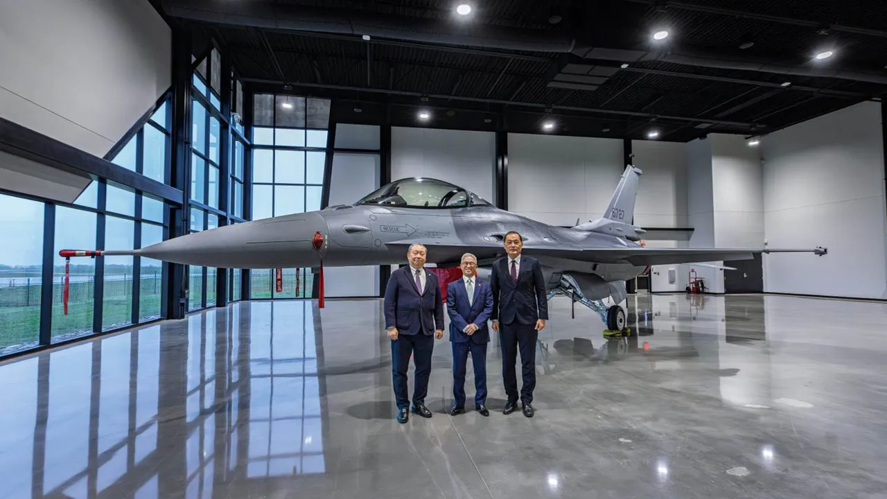 Three officials stand in front of an assembled gray F-16 Block 70 inside a production hangar.