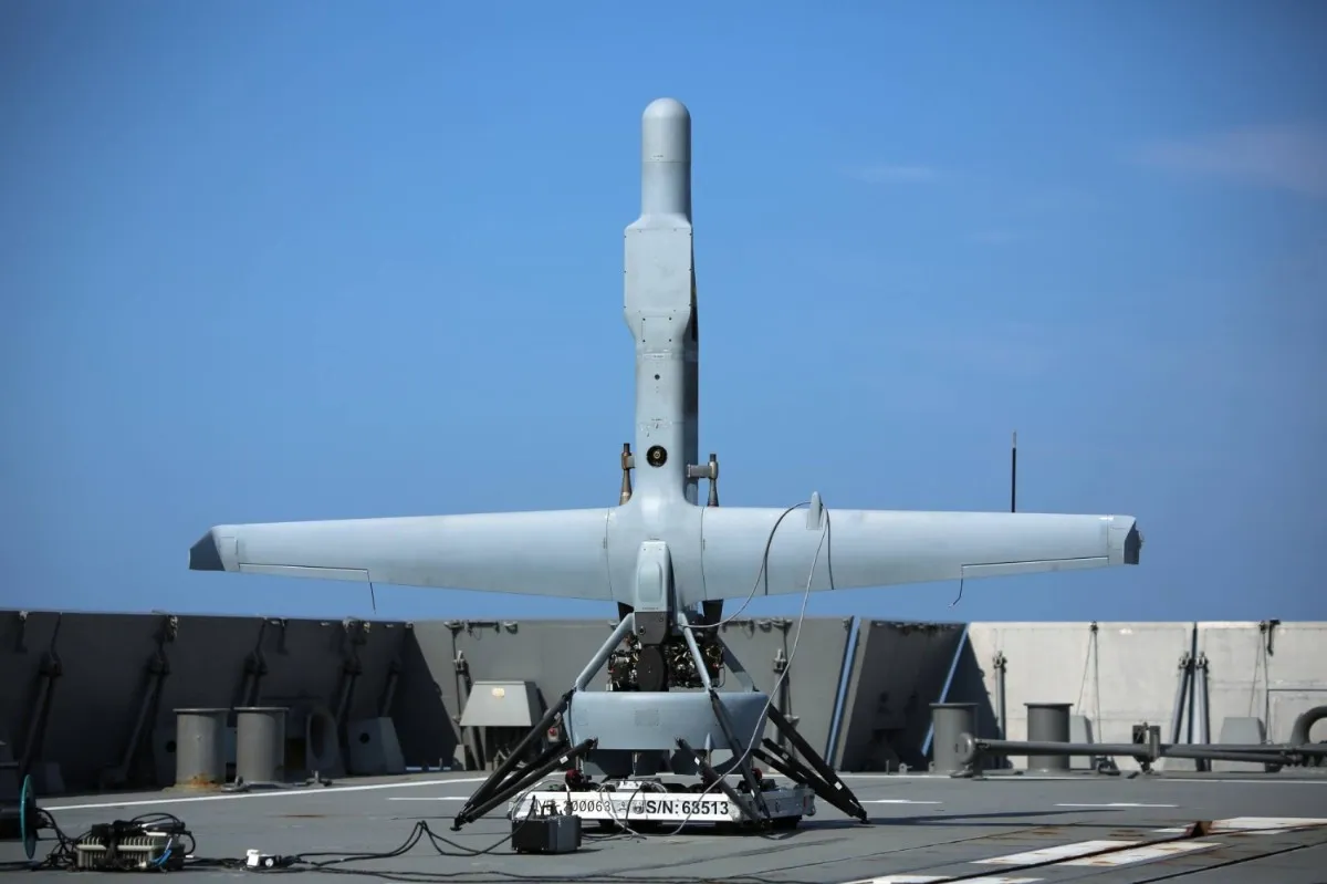 A grey V-BAT tail-sitter drone stands upright on its launch stand on a warship flight deck under a blue sky.