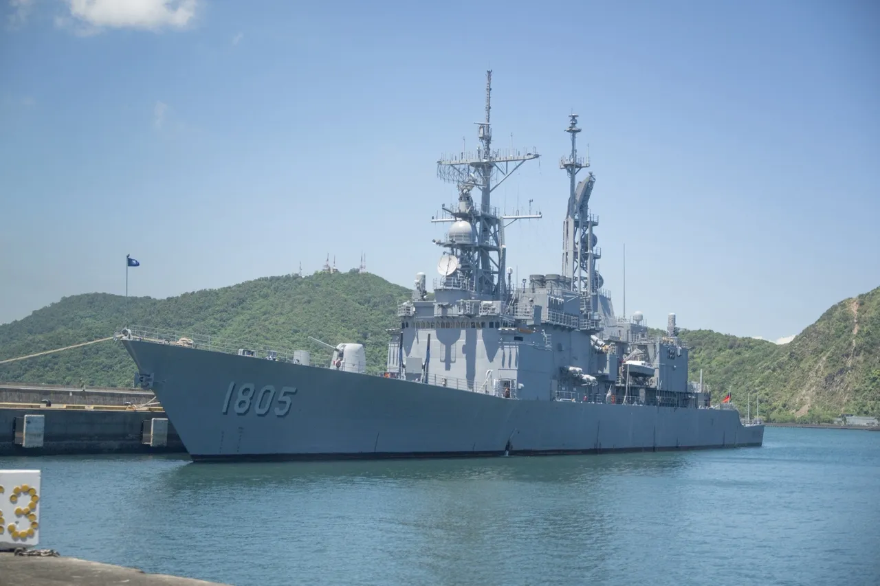 A grey navy destroyer with hull number 1805 moored at a concrete pier in calm blue water, with tall masts and radar domes and green hills behind it.