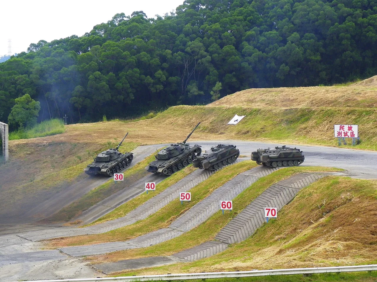 Two olive tanks and two tracked armoured carriers climb concrete test ramps beside a wooded hill