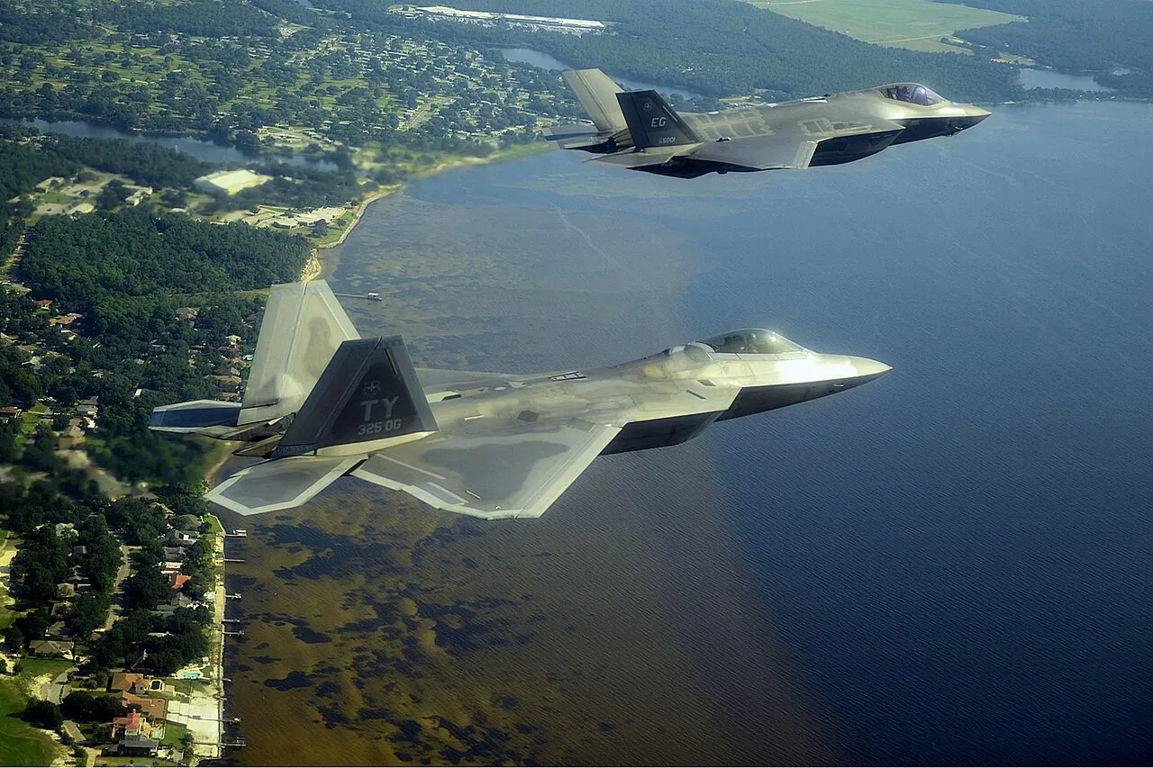 A grey F-22 stealth fighter in the foreground and an F-35 stealth fighter behind it flying over a coastline