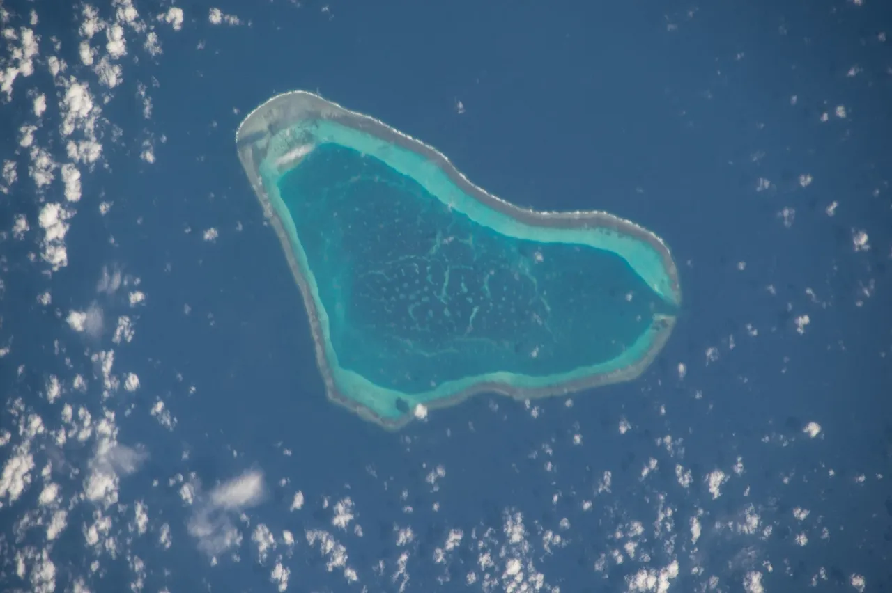 Aerial view from space of a triangular coral atoll with a turquoise lagoon surrounded by deep blue sea and scattered clouds