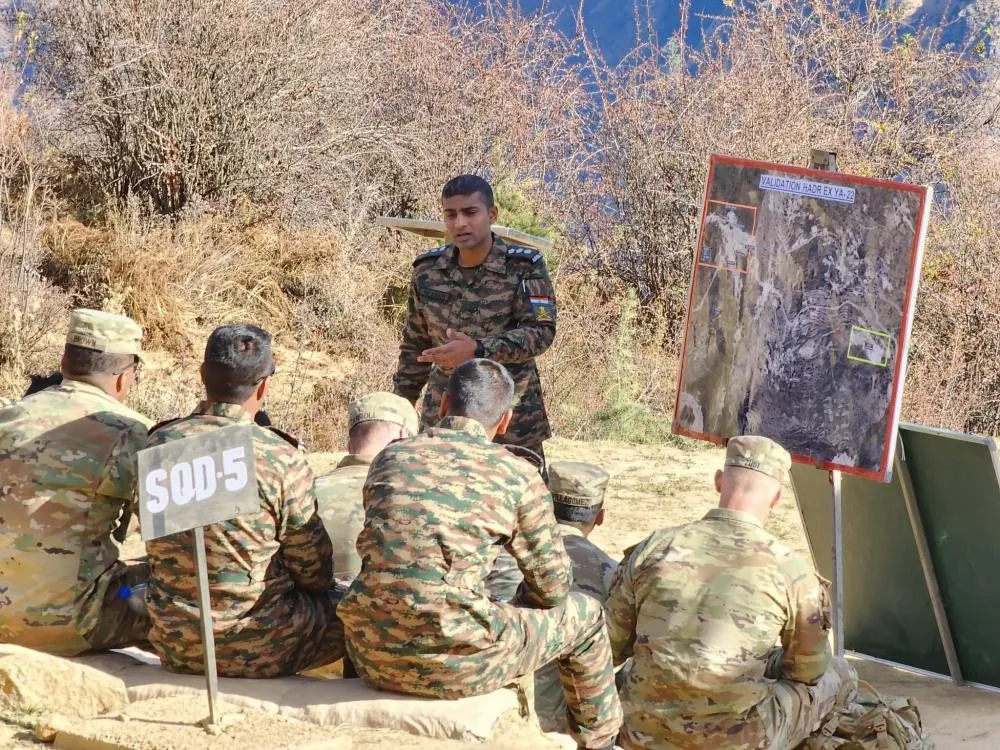 An Indian Army soldier in camouflage stands and speaks to seated Indian and US soldiers beside a map board on a dry hillside