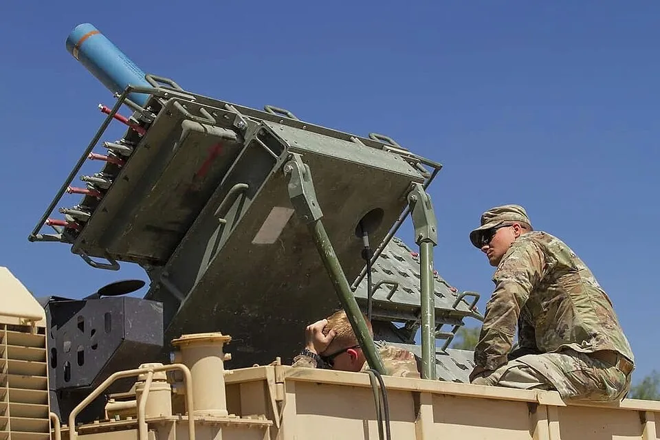 Two soldiers in camouflage uniforms sit on the bed of a tan military truck beside a large olive-green mine dispenser rack angled toward a clear blue sky, with a blue practice canister loaded at the top.