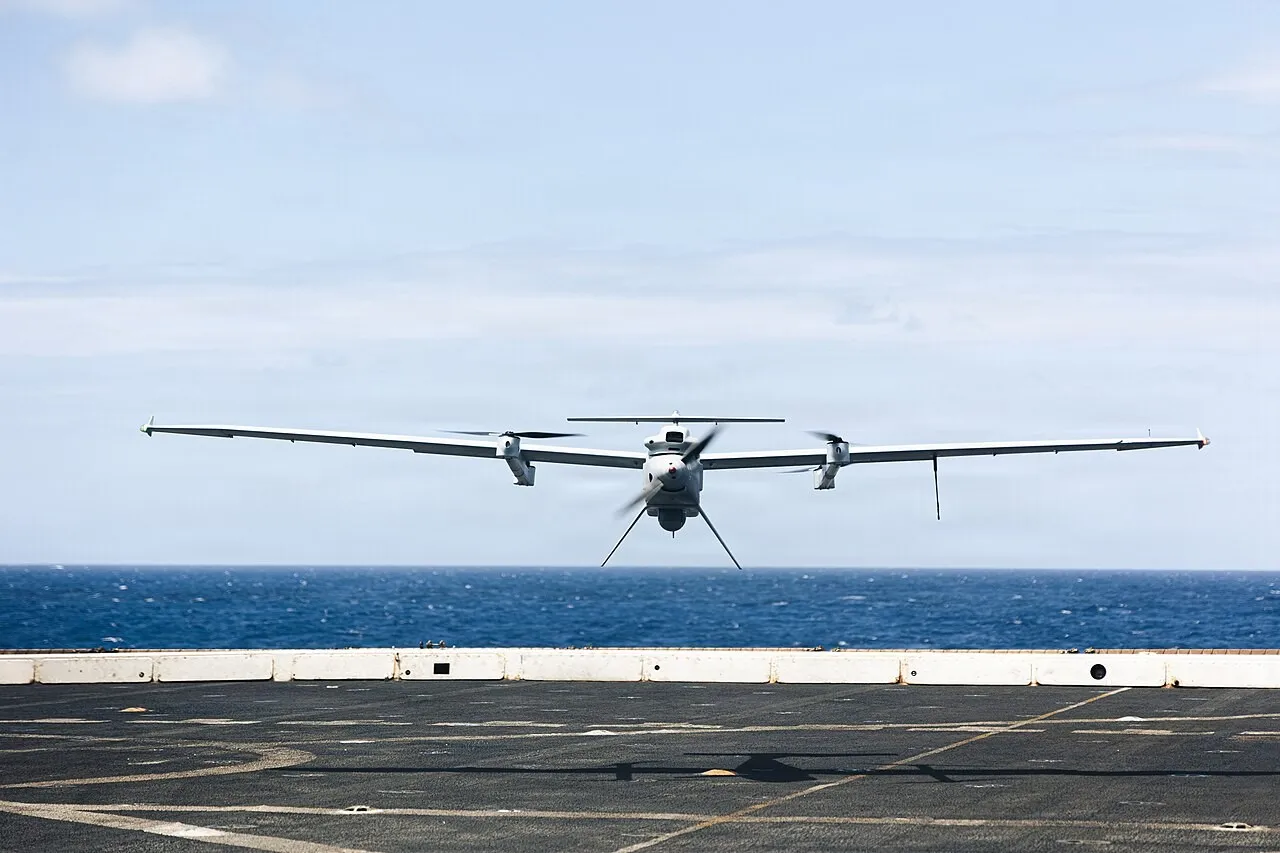 A white fixed-wing drone with long slender wings and four lift rotors hovers low over the grey flight deck of a ship, with blue sea and sky behind.