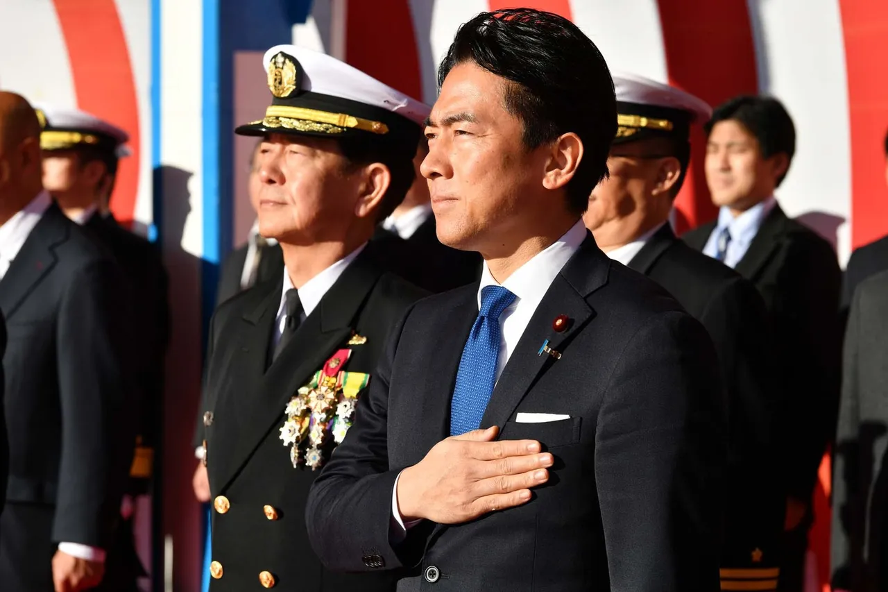 Japan's defence minister in a dark suit with his hand on his chest beside a uniformed navy admiral at a ship launch ceremony