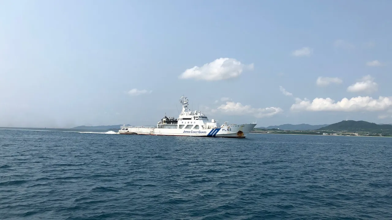 A white Japan Coast Guard patrol ship with a blue diagonal hull stripe sails on calm blue water with green hills on the horizon.