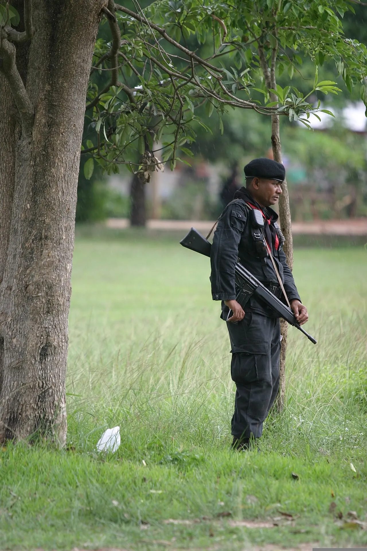 A Thai soldier in a black uniform and beret stands on grass beside a tree holding a rifle