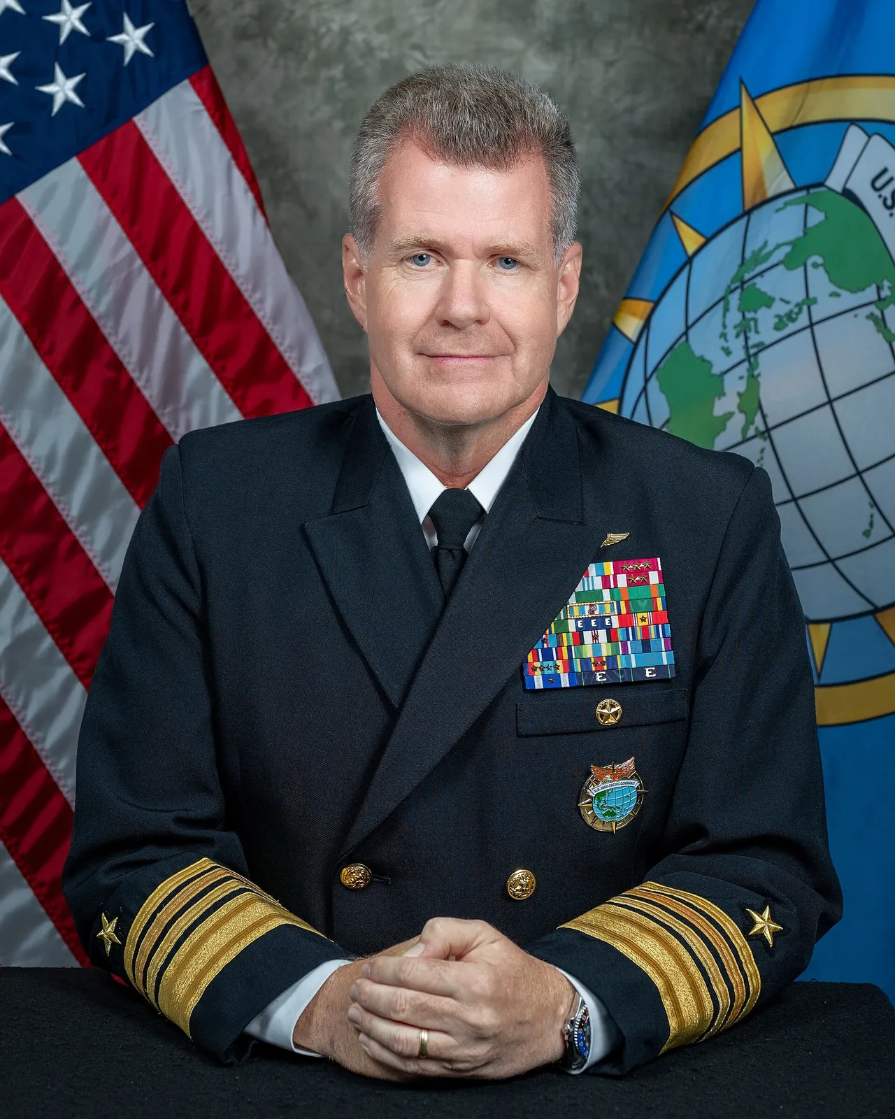 Official portrait of a grey-haired US Navy admiral in a dark dress uniform with medal ribbons, seated with hands folded in front of a US flag and a command flag