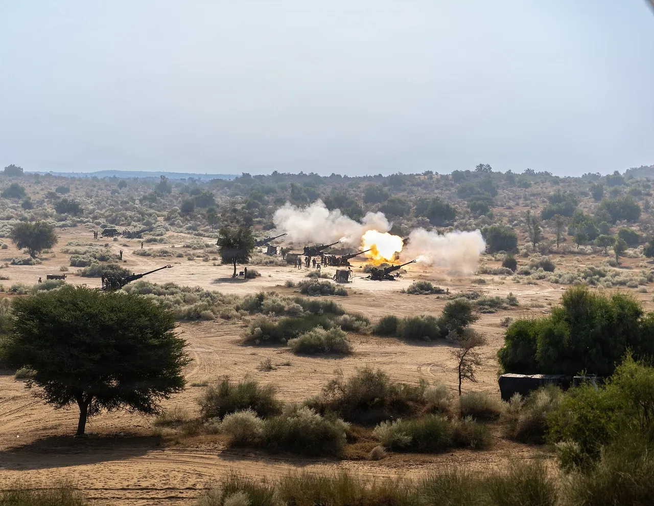Several towed howitzers firing with smoke and flame across a scrubby desert range