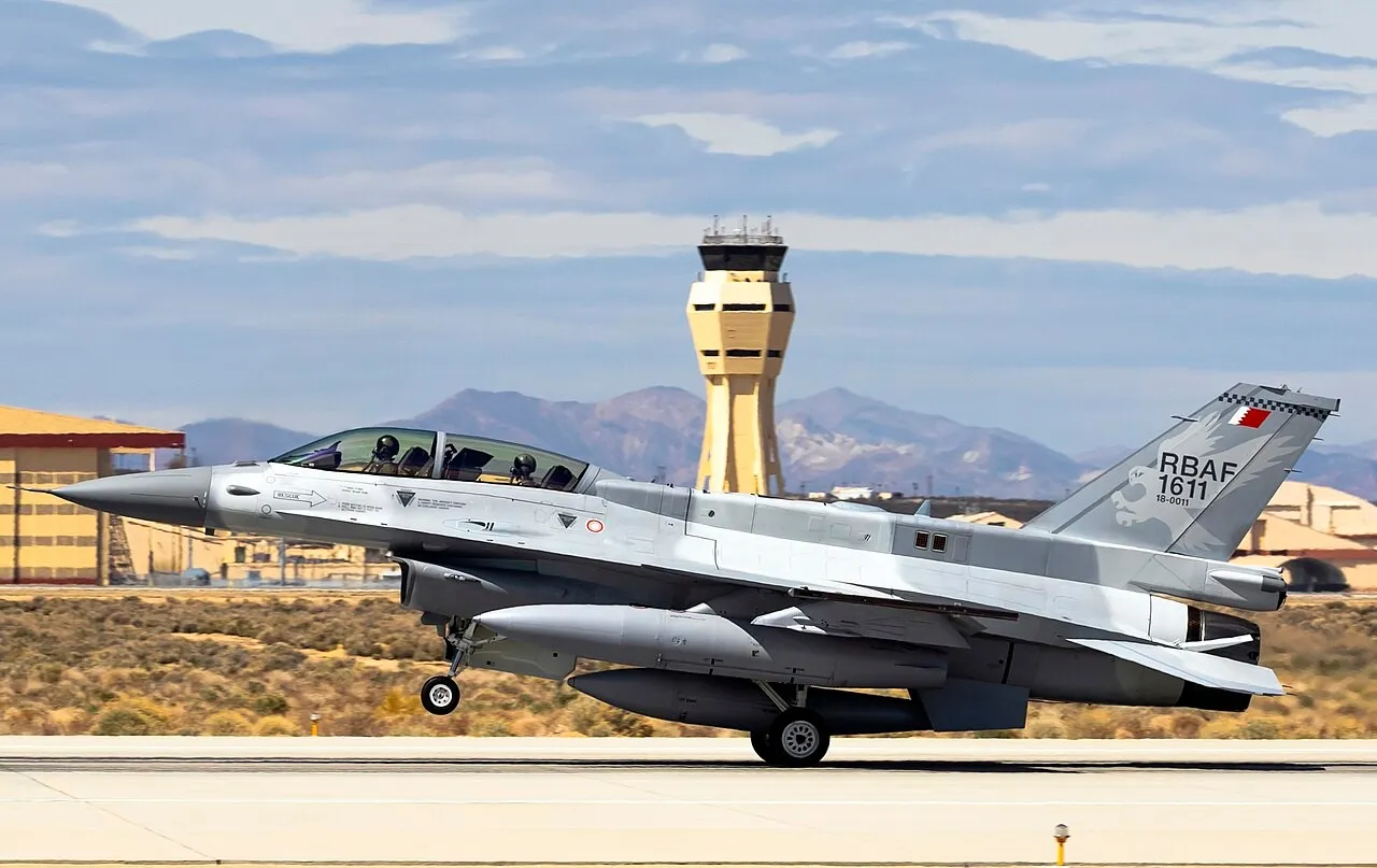 A grey two-seat F-16 fighter jet with Bahraini markings lands on a desert runway, with a yellow control tower and brown mountains behind it.