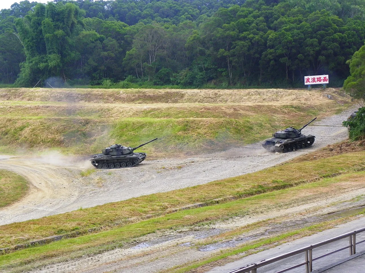 Two dark green M60A3 tanks drive along a dusty gravel track between grassy banks with a forested hill behind