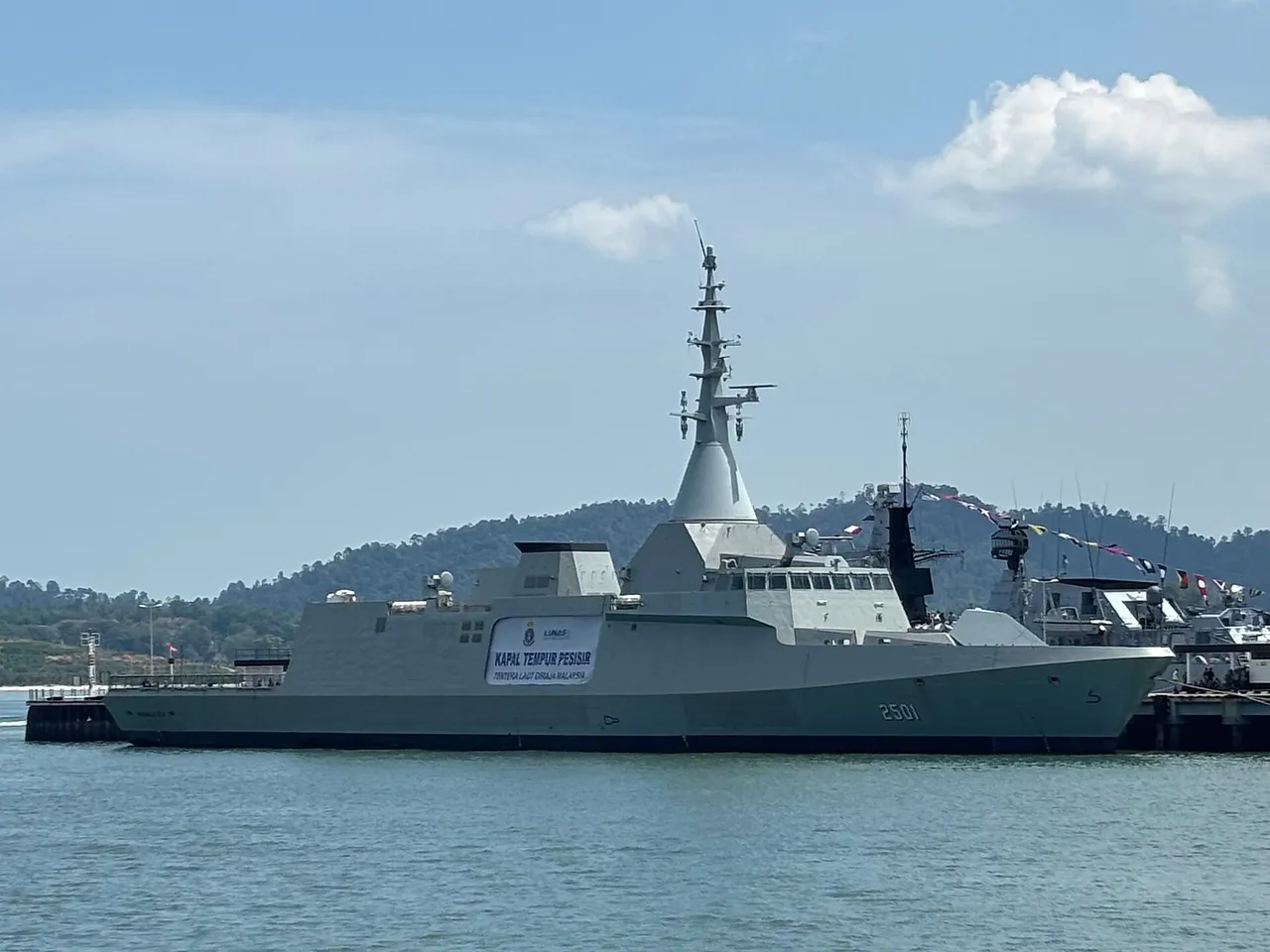 A grey stealthy littoral combat ship with a tall integrated mast and pennant number 2501 moored at a pier in calm water.