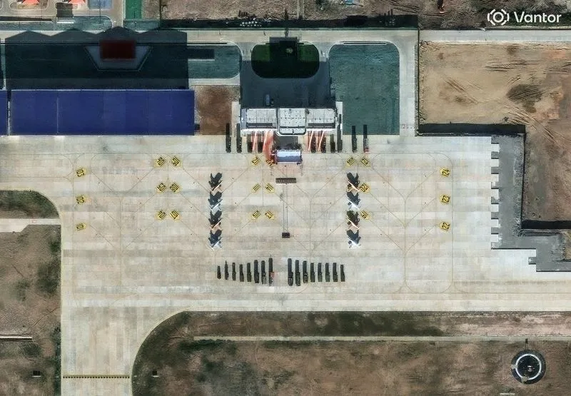 Overhead satellite view of an airport apron with eight small jets parked in two columns and a line of dark vehicles in front of a terminal building.