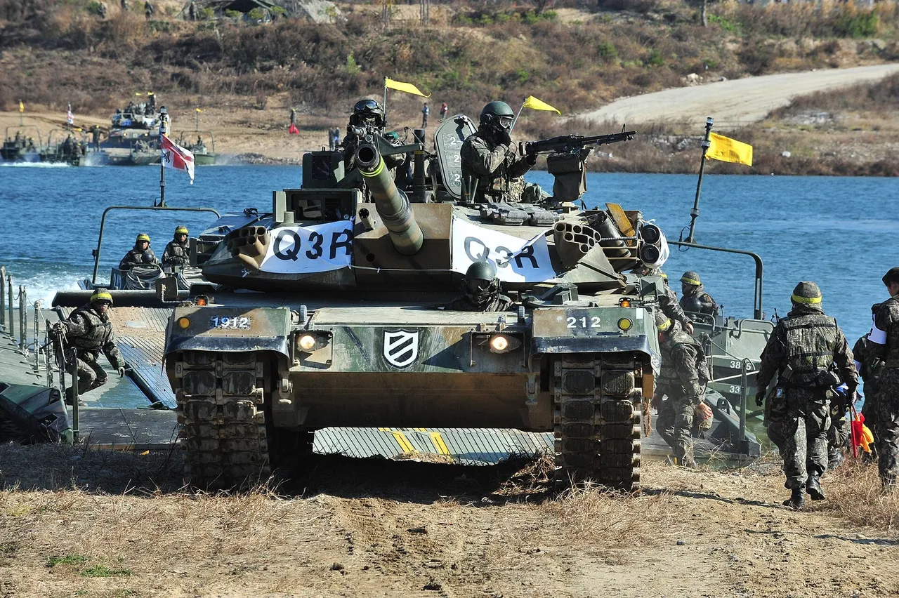 A South Korean army tank with crew in the hatches drives off a pontoon raft onto a riverbank as soldiers walk alongside