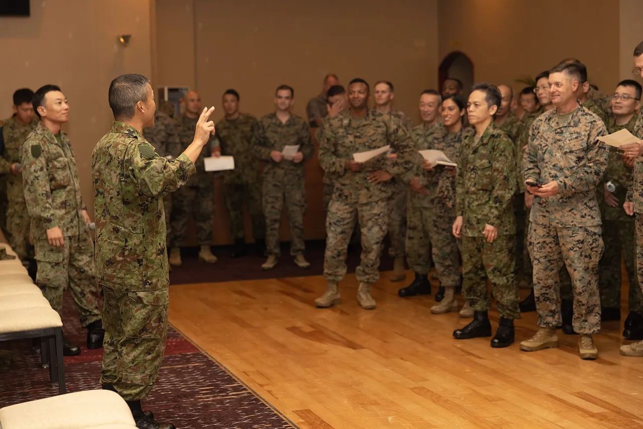 A Japanese soldier in green camouflage raises his hand as he leads a group of Japanese soldiers and US Marines standing in a hall
