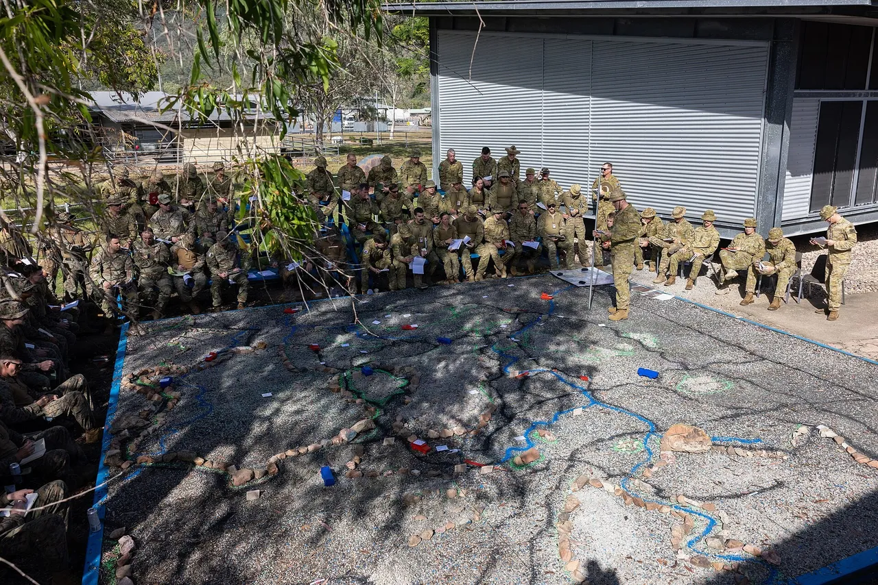 Rows of soldiers in camouflage uniforms seated around a large terrain model on the ground while an Australian soldier gives a briefing
