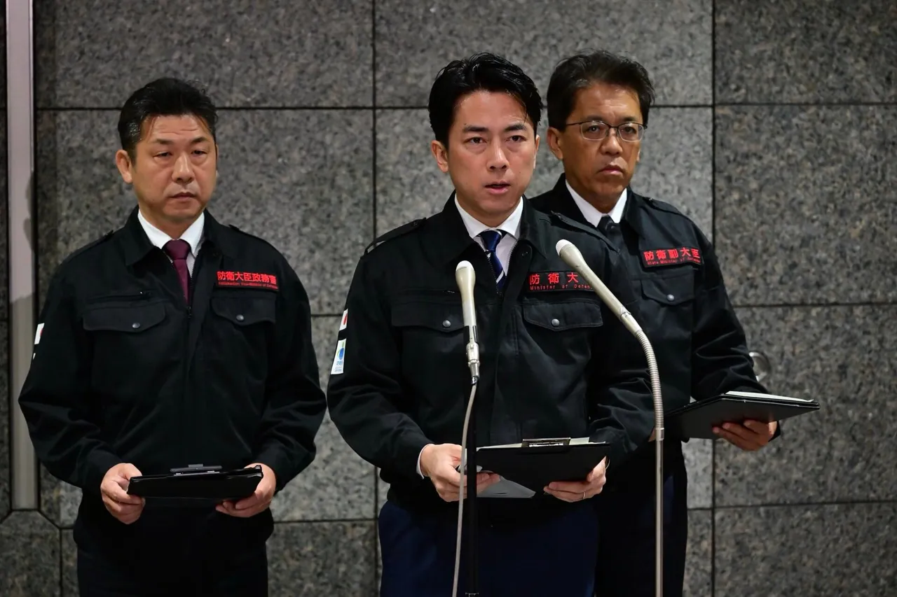 Japanese defence minister Shinjiro Koizumi in a dark work jacket speaking into microphones, flanked by two officials holding folders