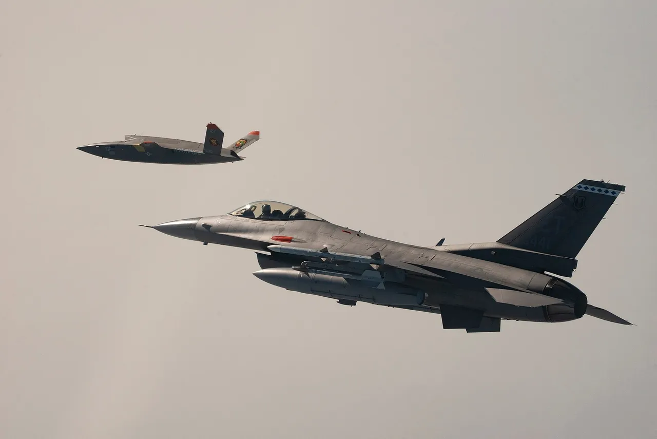 A grey drone with a V-shaped tail flies above and behind a grey F-16 fighter jet against a pale sky