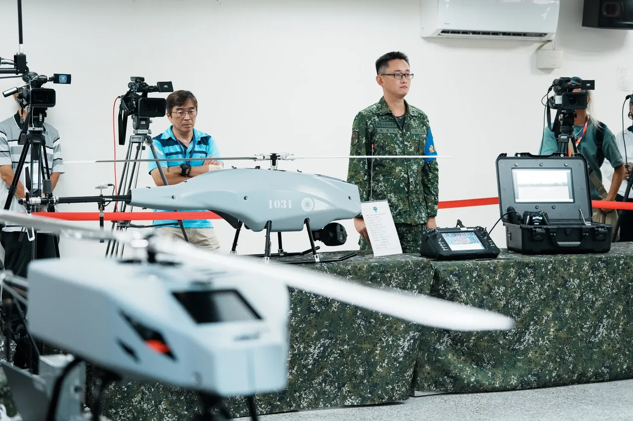 A Taiwanese soldier stands behind a table displaying a camouflaged helicopter drone and a fixed-wing drone, with camera crews in the background