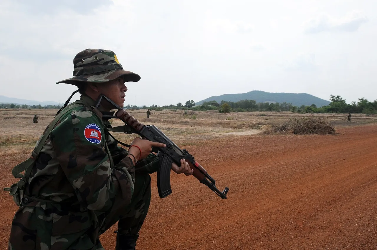 Cambodian soldier in camouflage and a bush hat holding a rifle on a red dirt road in open country
