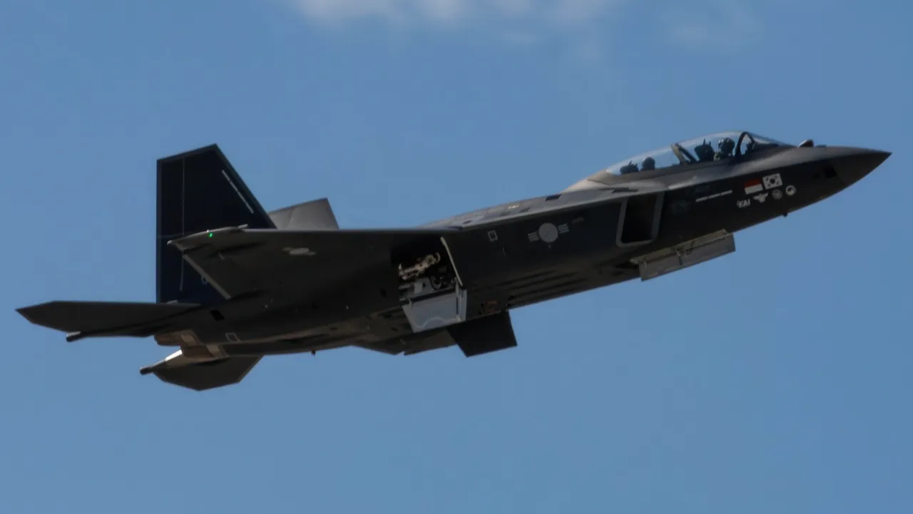 A dark grey twin-seat KF-21 fighter jet with South Korean markings climbs against a clear blue sky with its landing gear doors open