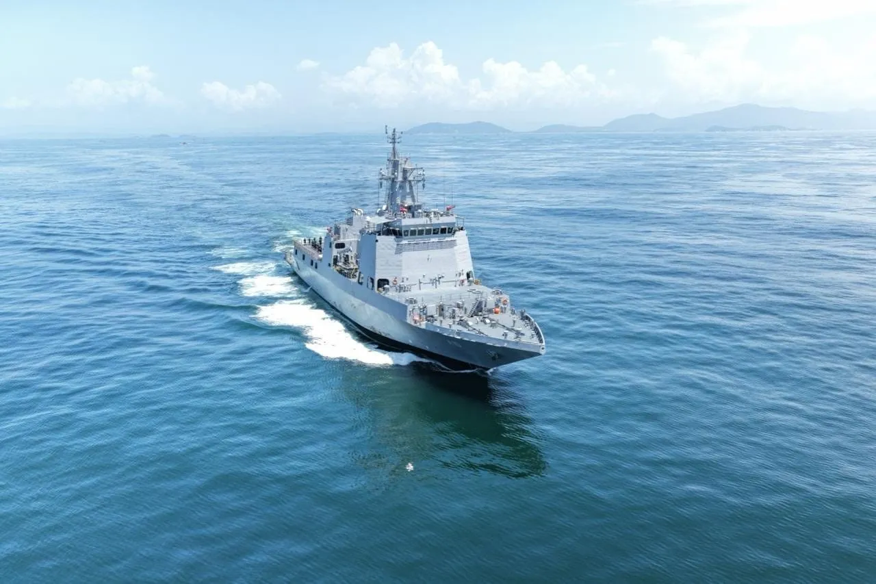 A grey naval vessel with a tall mast and a wide flat aft deck cuts through calm blue water, with hills on the horizon