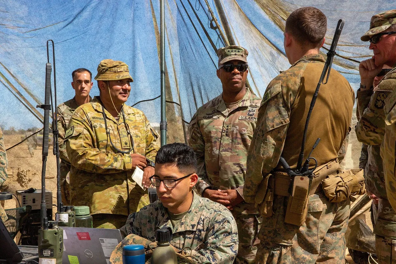 A US Army general in camouflage uniform and sunglasses stands in a field tent beside an Australian officer while a Marine works at a laptop