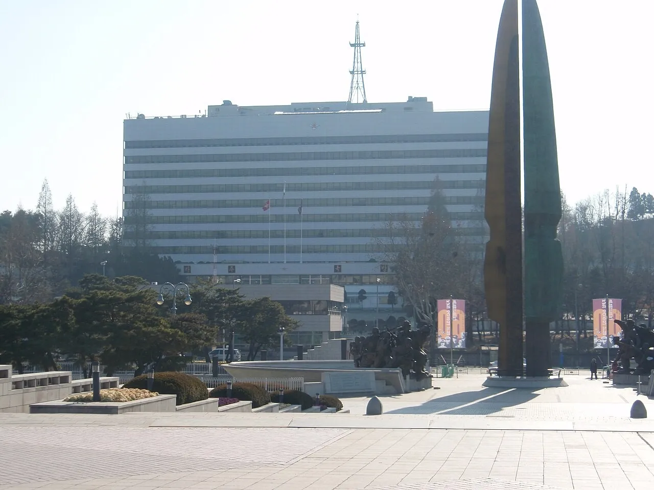 A large white multi-storey office building with a radio mast on its roof, seen across a paved plaza with a tall bronze monument and pine trees.