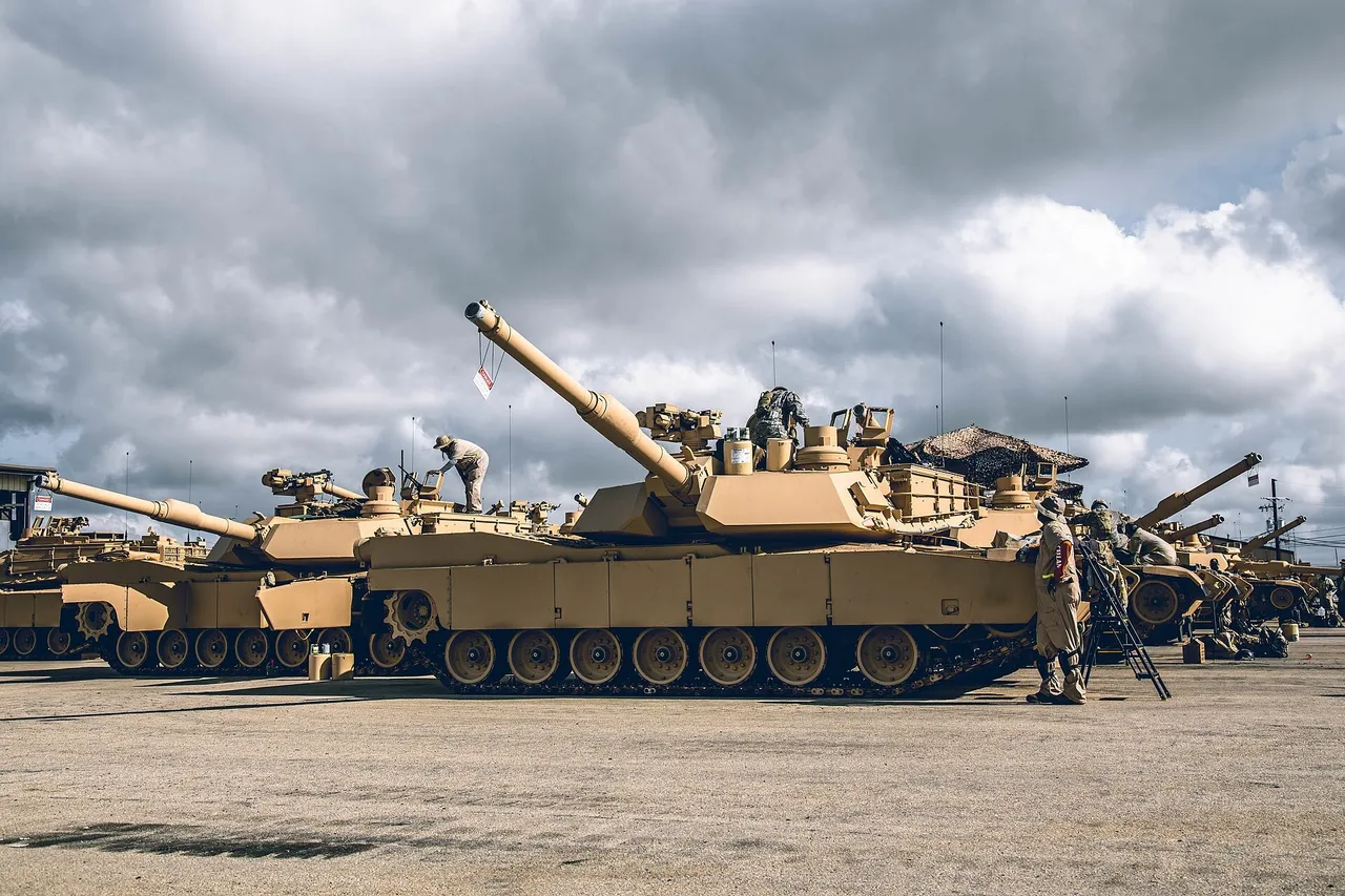 Desert-tan M1A2 Abrams tanks parked in a row on a gravel lot under a cloudy sky as soldiers work on their turrets