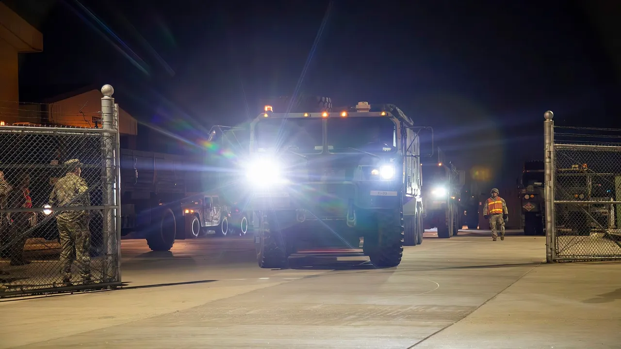 US Army trucks with headlights on drive out of a fenced gate at night during a convoy exercise in South Korea as a soldier watches
