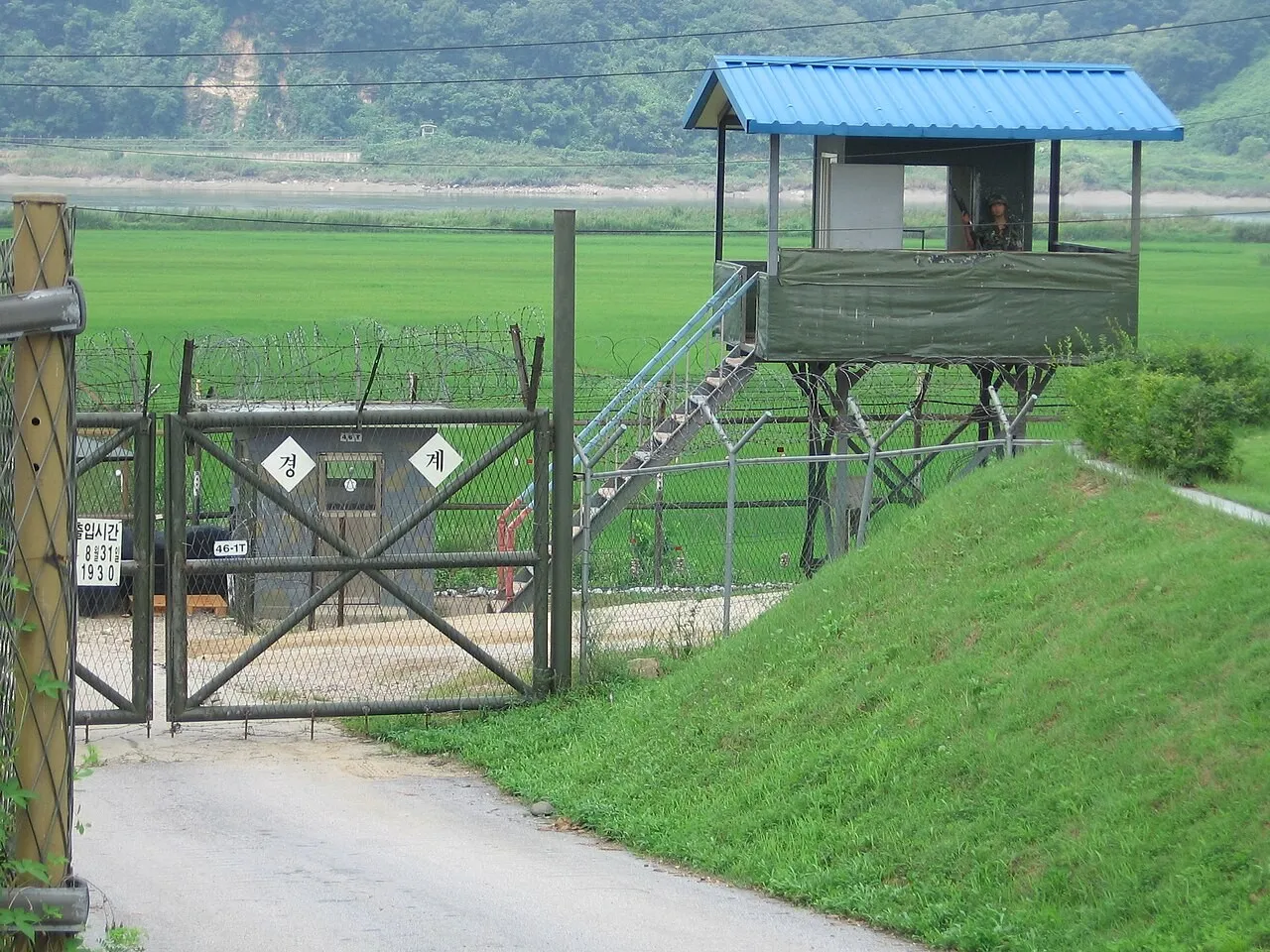 A South Korean soldier stands in a raised guard post with a blue roof beside a barbed-wire fence and gate, with green fields and a river behind.