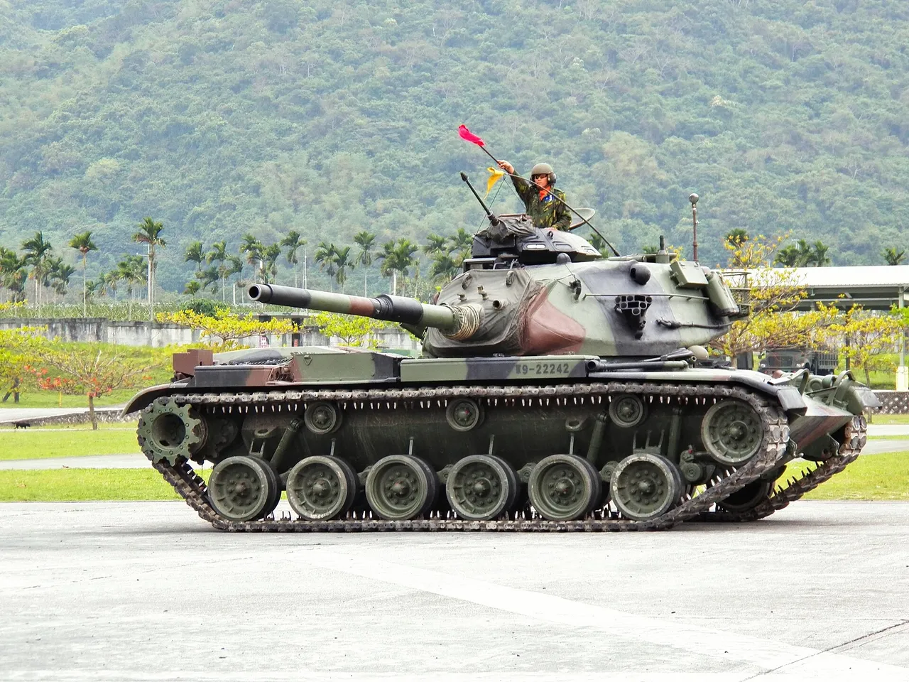 A camouflaged M60A3 tank with a crewman waving a red flag from the hatch drives across a parade ground with green hills behind