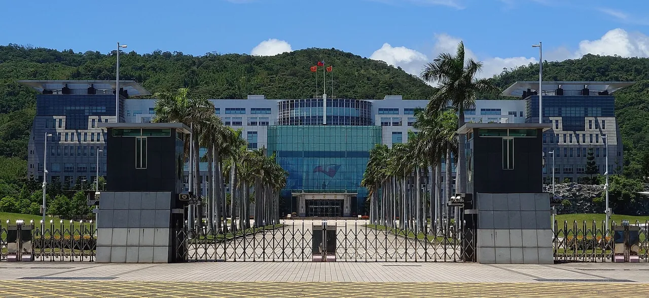 The glass-fronted Ministry of National Defense headquarters behind palm trees and a closed gate, with green hills behind