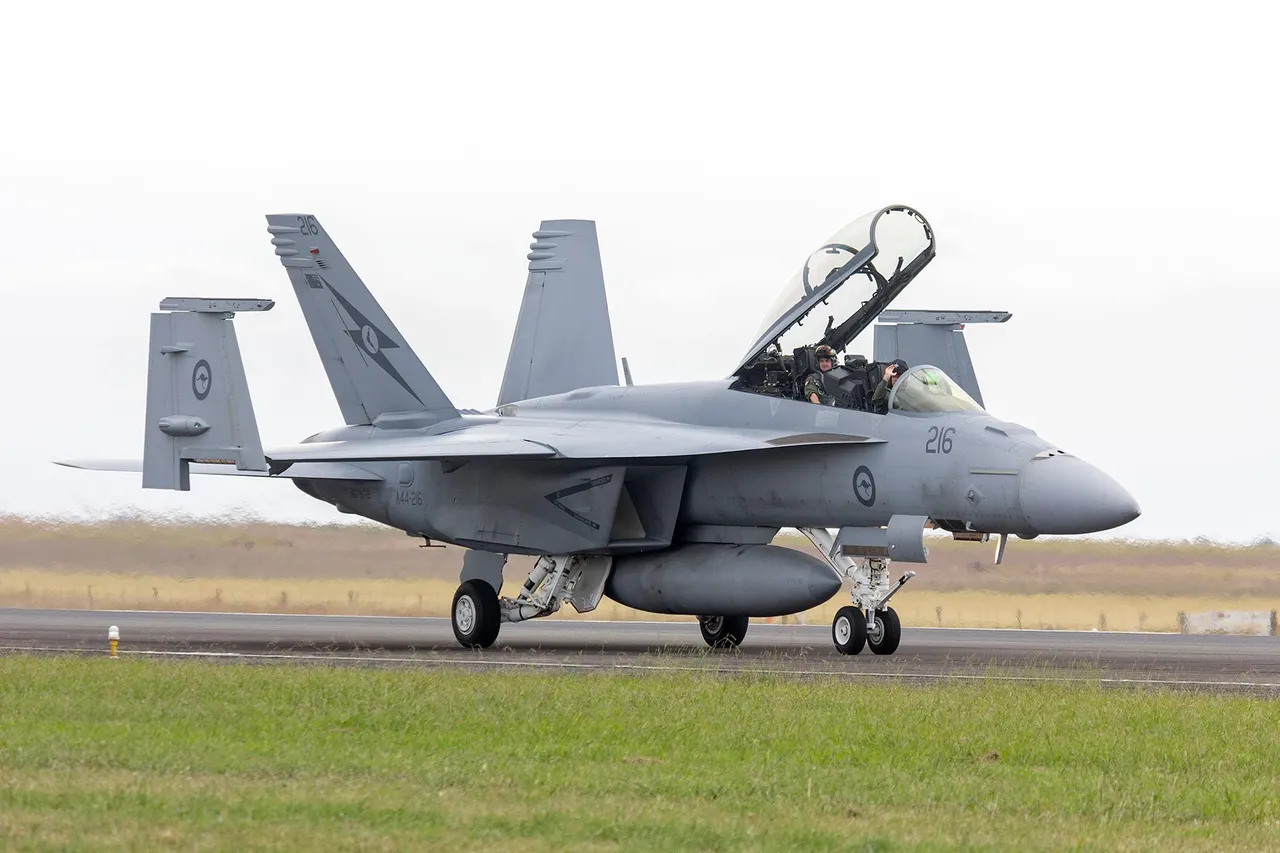 A grey twin-tail F/A-18F Super Hornet with its canopy open and two crew aboard taxis on a runway beside grass.