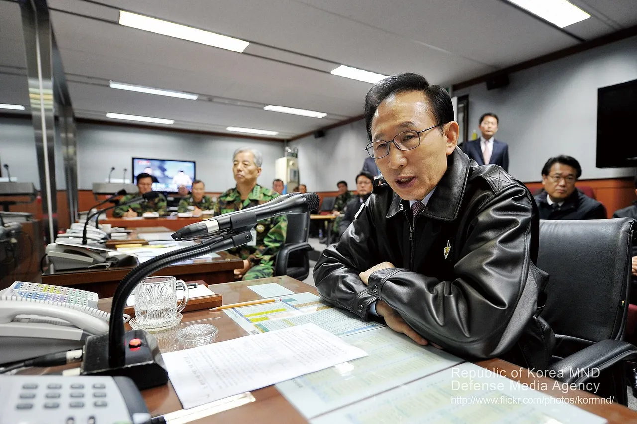 A man in a dark leather jacket speaks into a desk microphone in a military command room, with uniformed South Korean officers, telephones and screens behind him